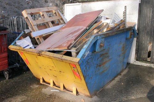 A large metal skip with a yellow front panel and blue sides, positioned on an asphalt surface in an outdoor area. The skip is filled with mixed construction and renovation debris, including wooden pallets, broken pieces of wood, a rusted sheet of metal, and various small fragments of building materials. The surrounding environment features a concrete wall behind the skip and part of a wooden fence to the right, with some metal objects visible on the left side. The debris appears to be haphazardly stacked, with some materials protruding over the edges of the skip, indicating recent disposal activity. The scene suggests an typical site in Muswell Hill where commercial waste removal services could be employed by companies such as Commercial Waste Removal Muswell Hill to clear construction or renovation refuse efficiently, ensuring the area remains tidy and compliant with local disposal regulations.