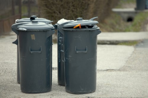 Two large dark grey plastic wheelie bins with closed lids are positioned side by side on an outdoor paved surface, likely a driveway or street in a residential or commercial area. The left bin has a uniform surface with a small badge or label on its lid, while the right bin’s lid is slightly open, revealing an orange handle and some visible rubbish inside. The background includes an out-of-focus natural environment with trees and shrubbery, and a blurred structure, possibly a bridge or tunnel, in the distance. The scene is lit with natural daylight, suggesting a typical day in Muswell Hill, London. The image relates to waste disposal and rubbish removal services, aligning with offerings from Commercial Waste Removal Muswell Hill, which caters to local waste management needs and maintains clear, organized waste collection practices.
