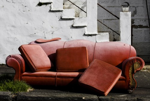 An outdoor scene featuring a worn, terra cotta-colored sofa with matching cushions and an ottoman, placed on a paved surface adjacent to a concrete staircase with missing and cracked steps. The sofa's fabric appears faded and slightly weathered, with visible signs of wear. Small patches of grass and moss grow at the base of the furniture, indicating it has been discarded outdoors. In the background, a concrete wall and staircase with metal railing elements are visible, suggesting an urban or residential setting. The lighting is natural, casting soft shadows across the scene, and the overall environment reflects an area where rubbish, such as old household furniture, has been left for collection or removal, typical of waste removal services in Muswell Hill.