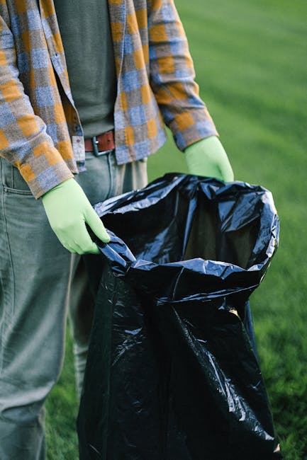A person wearing a mustard and grey checkered shirt over a dark green top, with light grey trousers secured by a brown belt, is standing outdoors on a grassy area. They are holding an open black plast