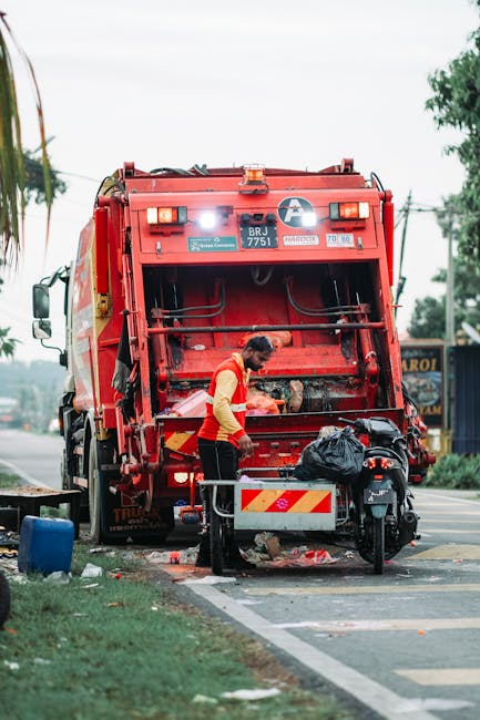 A waste collection worker operating a large red rubbish truck on a suburban street, seen from the rear with its compaction mechanism open. The worker, dressed in a high-visibility yellow and orange ve